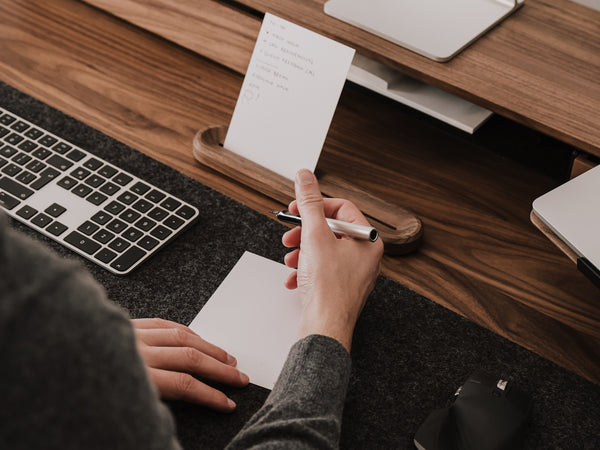 Person taking notes or tasks at a desk with a laptop and keyboard |-