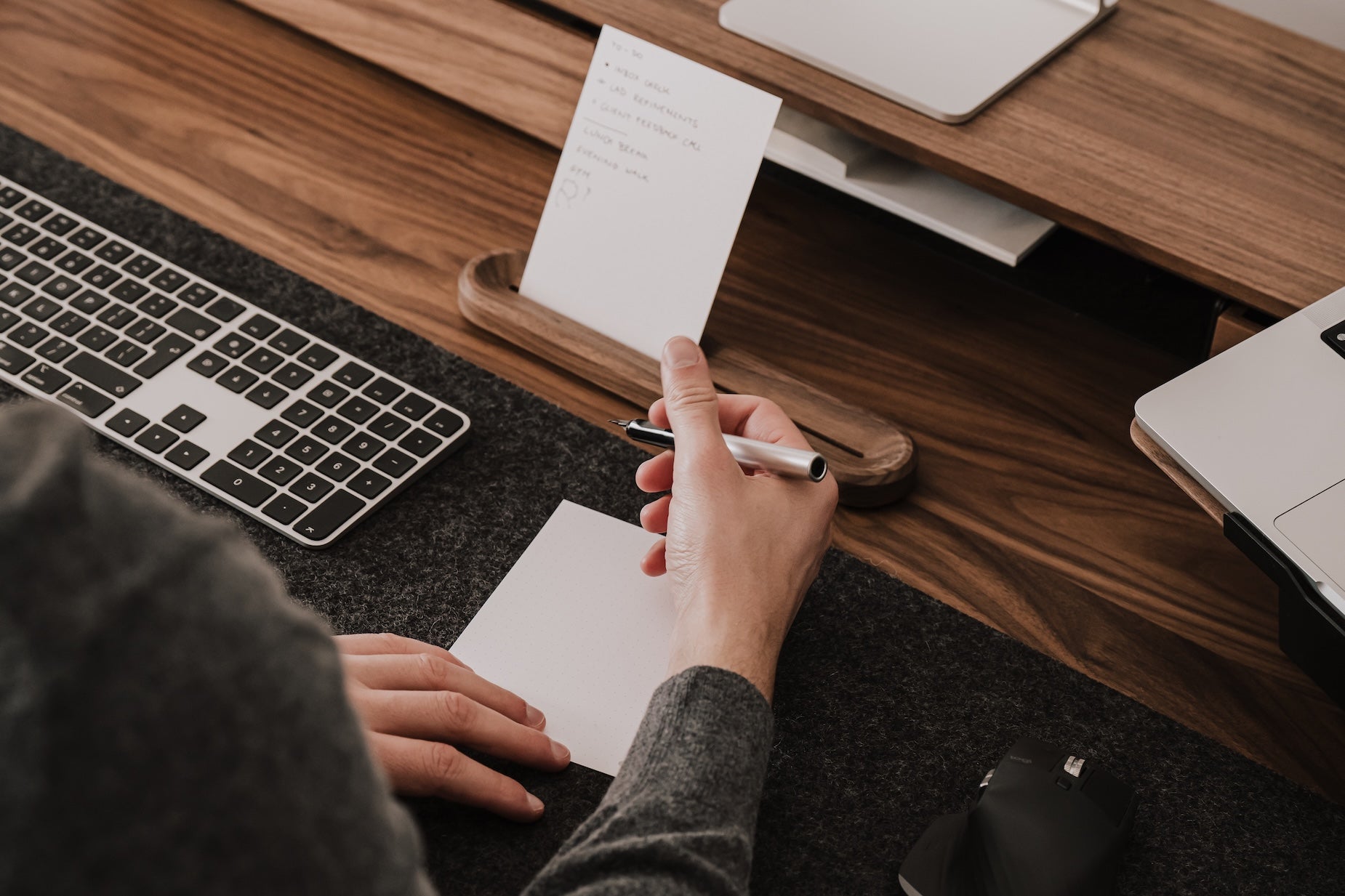 Person taking notes or tasks at a desk with a laptop and keyboard |-