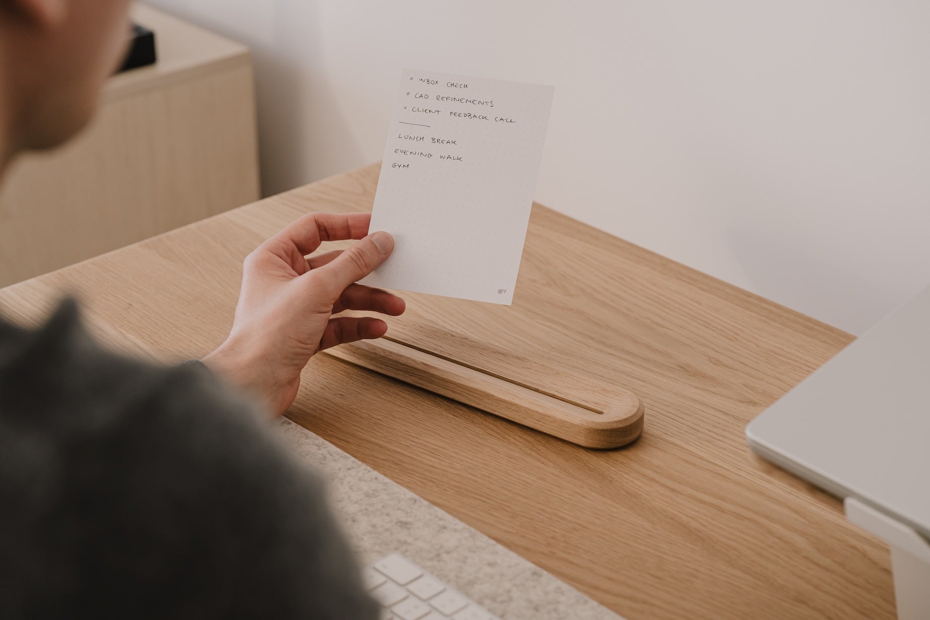 Person holding a paper to-do list at a wooden desk with a laptop in the background |-