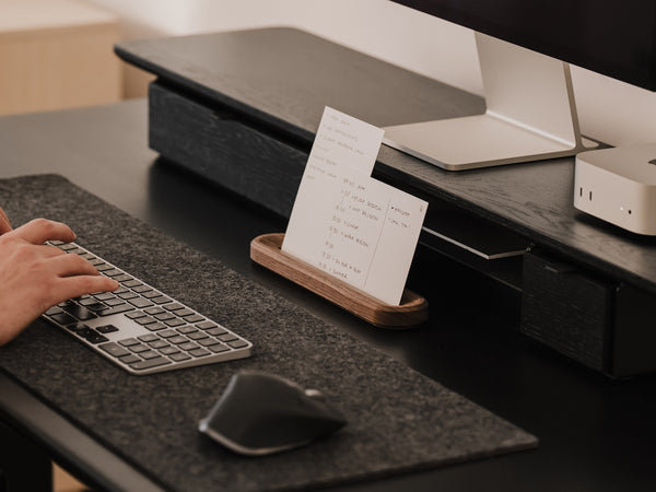 Person using a computer keyboard at a desk with a monitor and small stand holding a to-do list | walnut