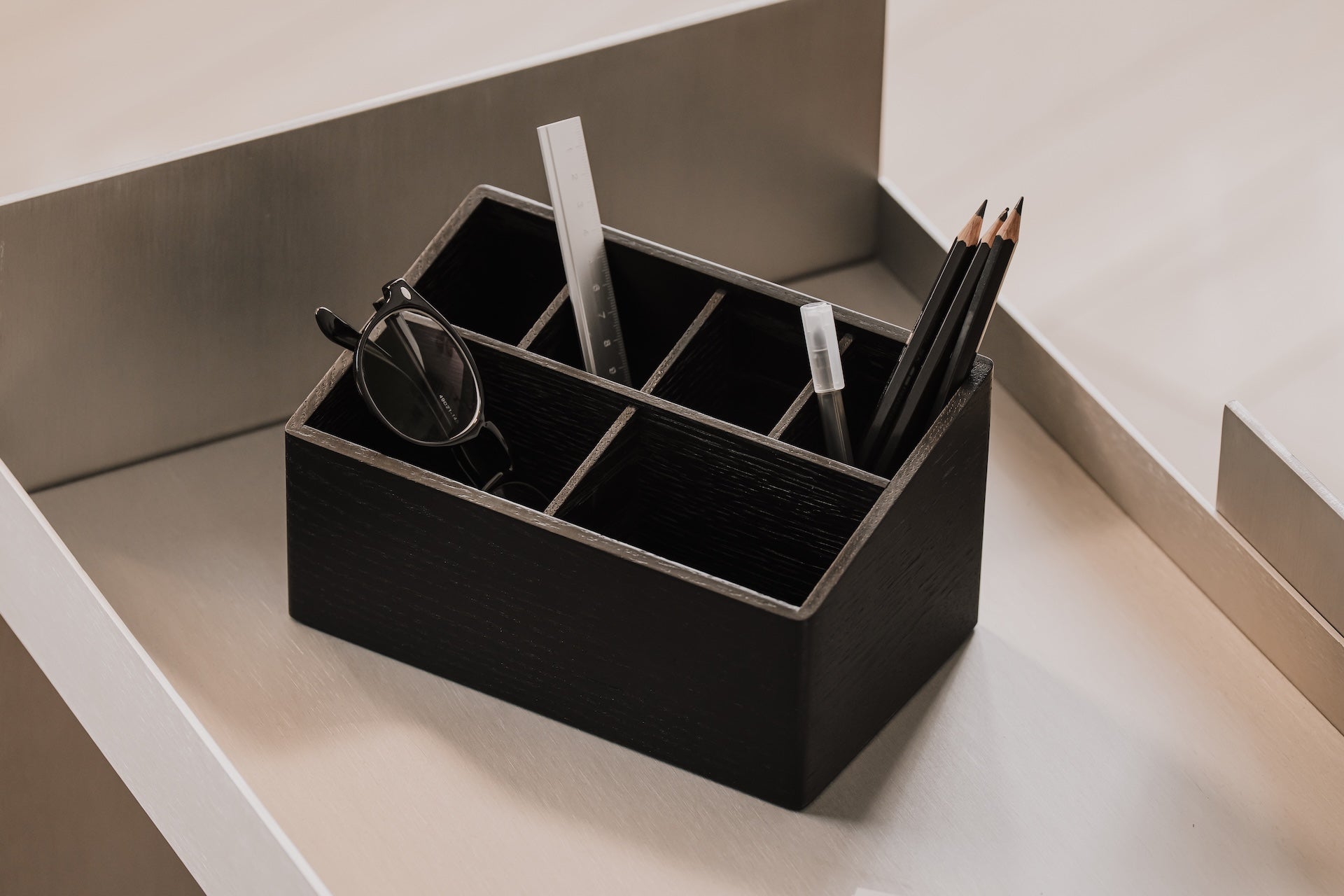 Black desk organizer with compartments organising glasses and pencils on a neutral stainless steel background | black