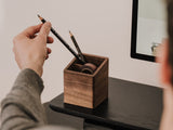 Person placing a pencil into a wooden pen holder on a desk. | walnut