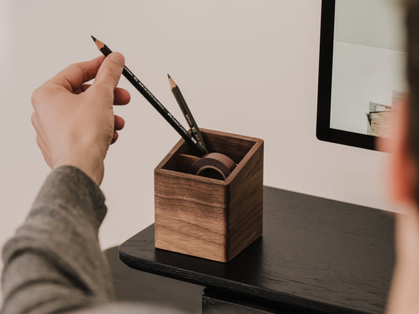 Person placing a pencil into a wooden pen holder on a desk. | walnut
