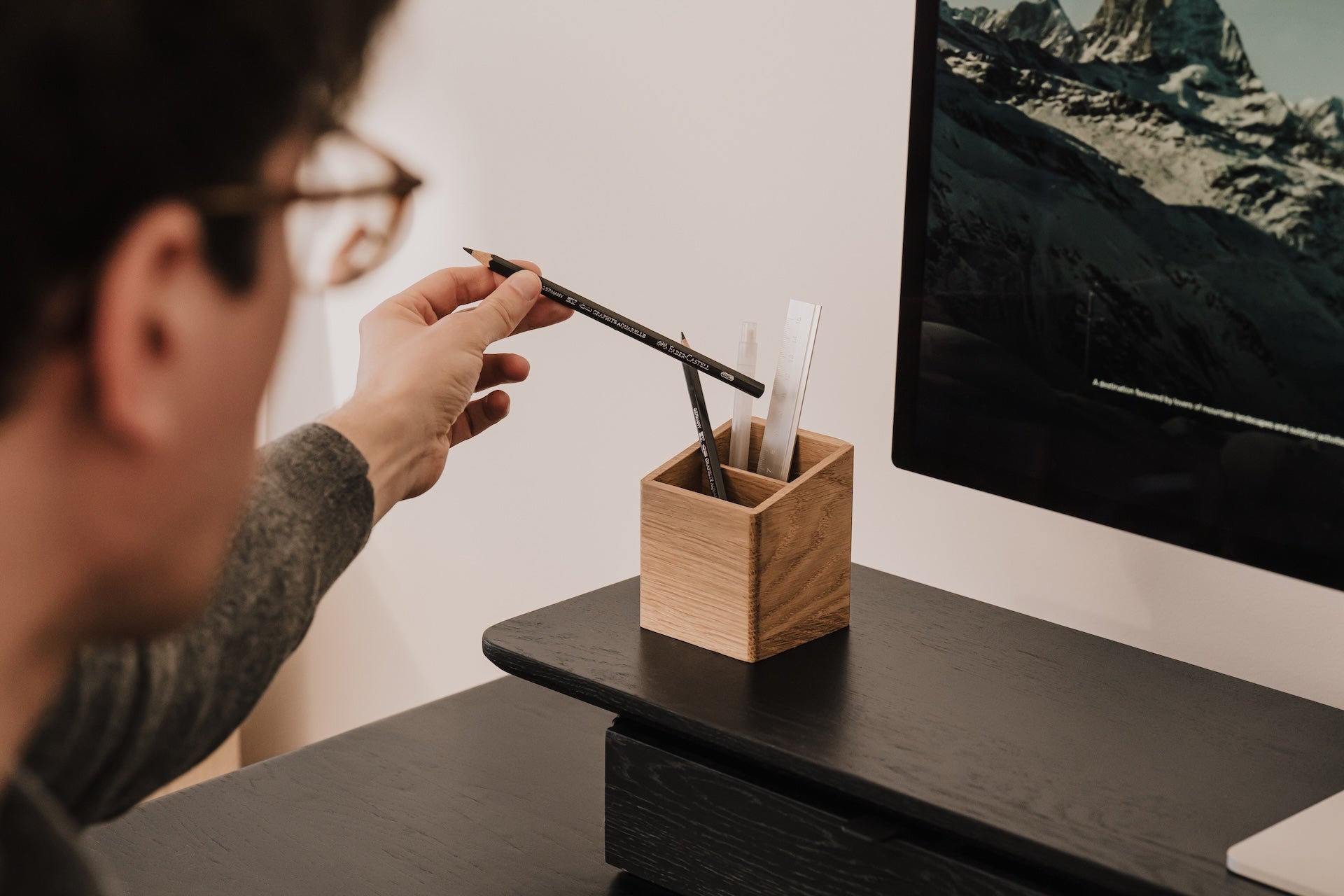 Person reaching for a pencil from a wooden pen holder on a desk in front of a computer screen. | oak